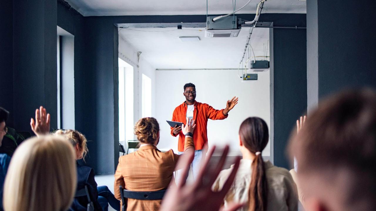 Handsome cheerful man in a orange shirt standing in front of an audience holding a tablet and using hand gestures to interact with the audience.